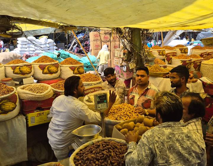 Bustling spice market stall in Delhi with vendors and sacks overflowing with nuts and dried fruit.