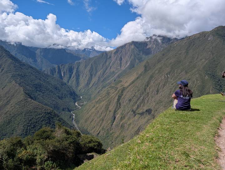 Traveler sits on grassy ledge admiring a vast green river valley framed by towering Andean peaks.