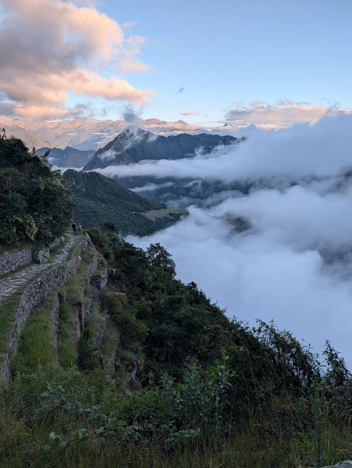 Stone Inca trail winds above clouds on a verdant mountainside in early light.