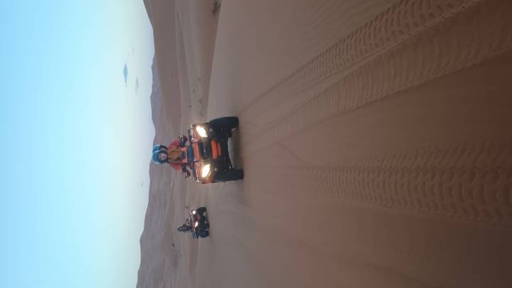 Two riders power quad bikes across soft desert sand leaving curving tyre tracks behind.