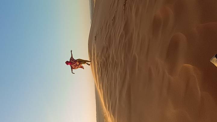 Traveller leaps joyfully on a wind-swept dune crest against a pale desert sky.