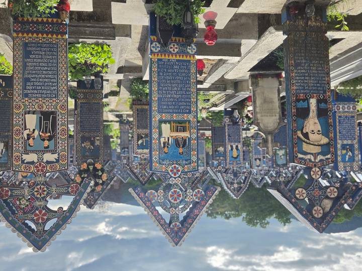 Rows of ornate blue and red carved crosses extend through the Merry Cemetery with hills behind.