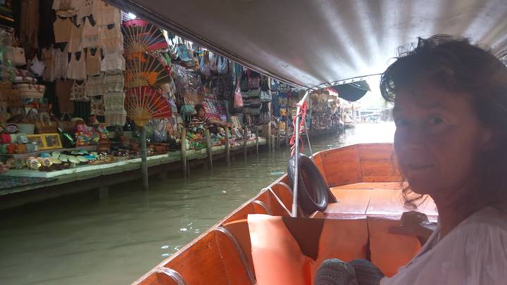Tourist seated in a long-tail boat passing stalls at a bustling Thai floating market canal.
