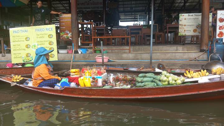 Vendor in a wooden boat selling fresh fruit and snacks at a floating market canal with restaurant stalls behind.