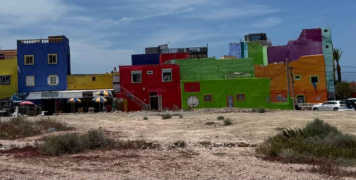 Row of brightly painted buildings in vivid blues, reds and greens bordering a sandy vacant lot under clear sky.