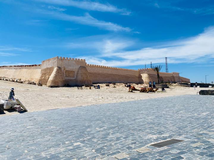 Historic stone ramparts of a seaside fortress under bright blue sky with scattered vendors nearby.