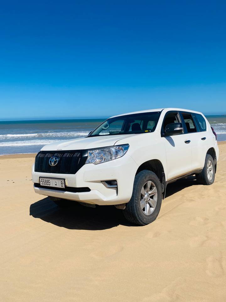 White Toyota 4x4 parked on a sandy beach beside gentle ocean waves and blue sky.