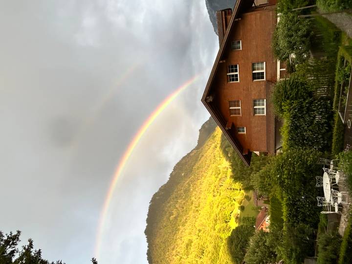 Double rainbow arching over an alpine valley with wooden chalet and green mountainside.