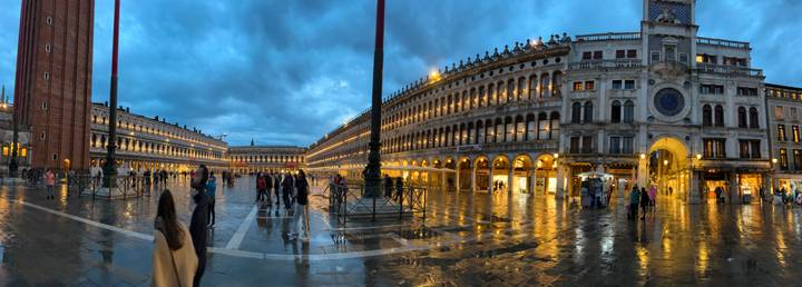 Evening panorama of wet Piazza San Marco with glowing arcades and visitors after rain.
