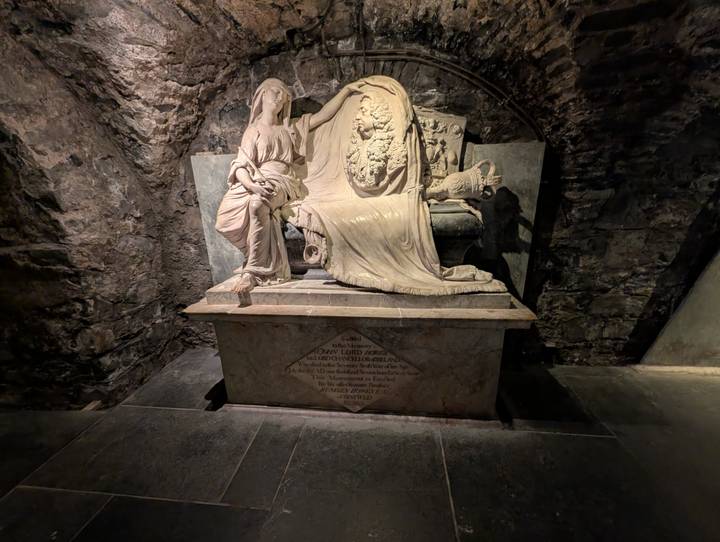 Marble funerary sculpture of a robed figure and coat of arms displayed inside a stone crypt.