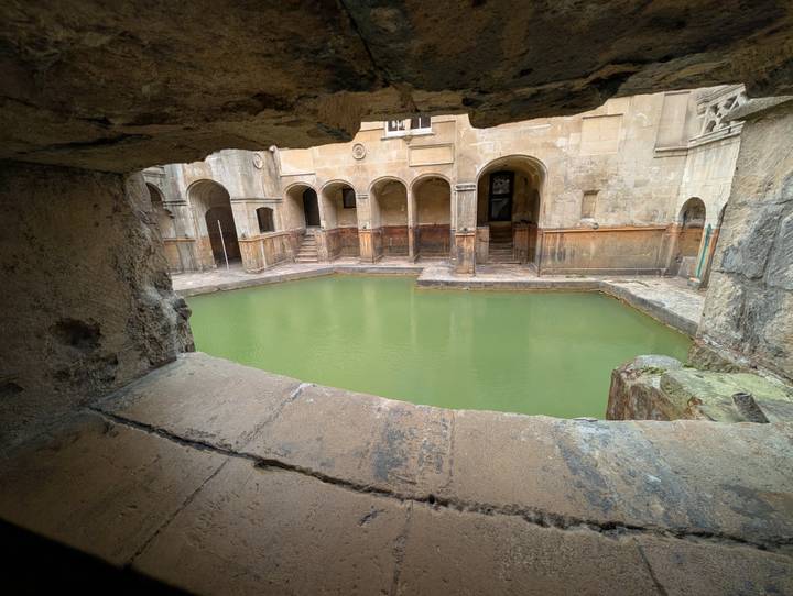 Historic Roman Baths pool filled with green water surrounded by weathered stone arches.