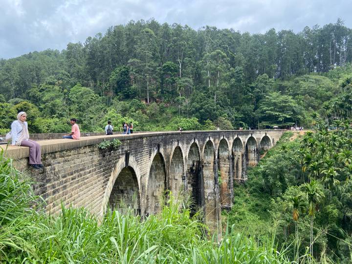 Historic nine-arch stone railway bridge spanning a deep forested gorge with visitors sitting along its edge