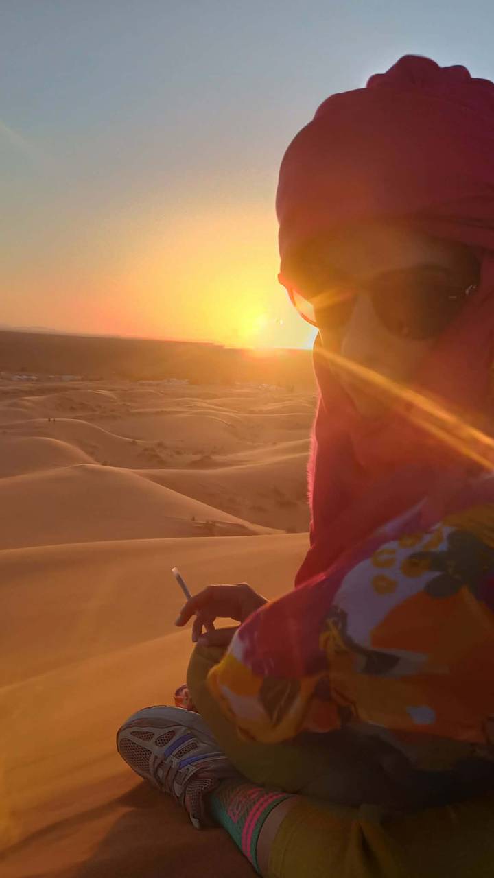 Close-up of a traveler in a pink scarf watching an orange sunset over endless sand dunes.