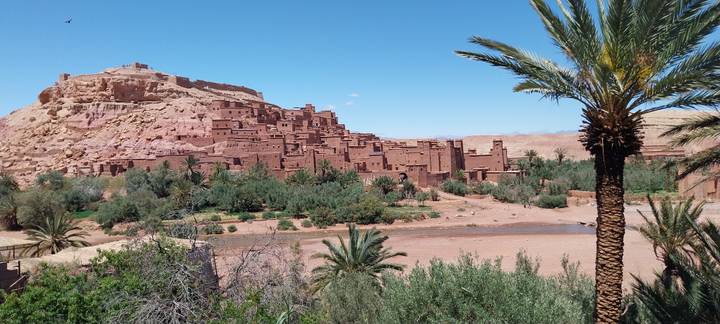 The mud-brick kasbah of Ait Benhaddou perched on a hill above a riverbed lined with palms.