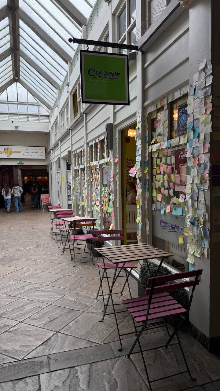 A cosy arcade where café tables face a shopfront plastered with colourful sticky notes and messages.