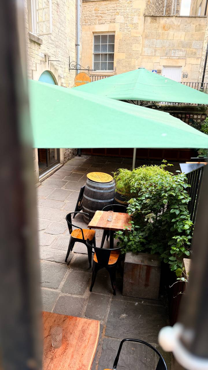 A small courtyard café with wooden barrel tables, greenery and a shaded umbrella-covered setting.