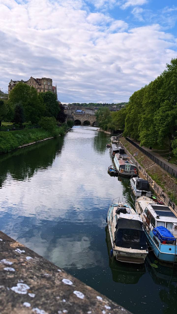 Calm river with moored narrowboats leads toward an arched stone bridge and historic Bath skyline.