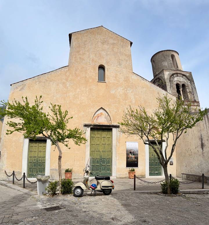 Simple stucco church façade with green wooden doors and young trees against a pale sky.