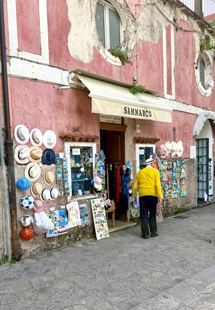 Small souvenir shop with hats and postcards adorns a pink plaster wall as an elderly customer browses.
