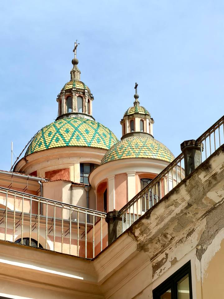 Colorful tiled domes of a Mediterranean church rise above peach-colored walls and railings against a clear blue sky.
