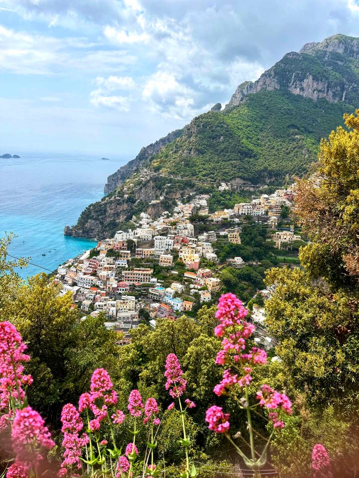 Panoramic postcard view of Positano’s pastel buildings cascading down to the bright blue Tyrrhenian Sea with pink flowers in the foreground.