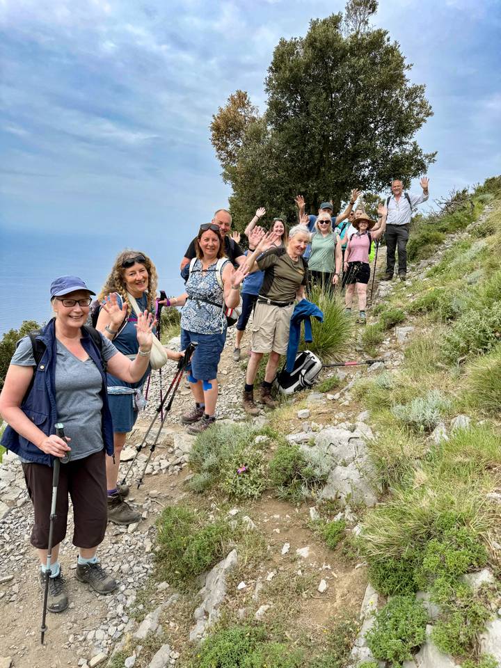 Smiling hiking group waving while trekking a rocky coastal trail high above the sea.