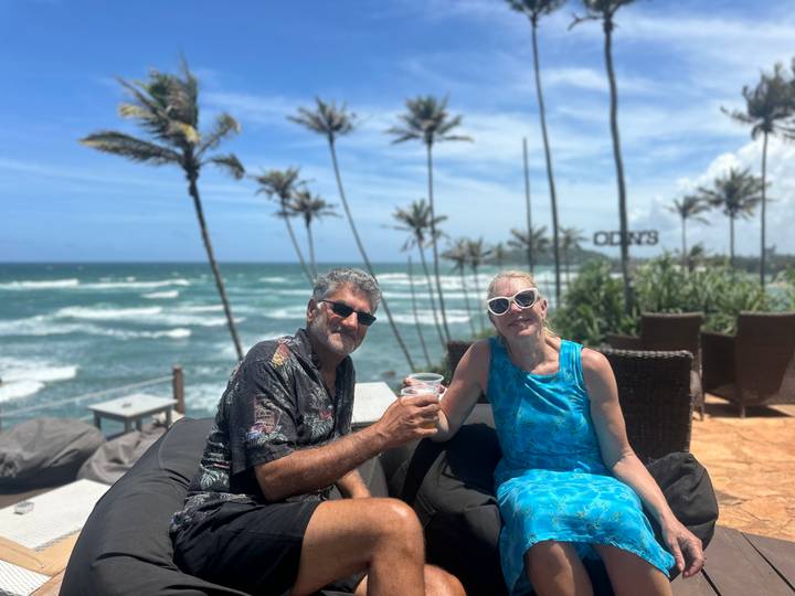 Couple toasts drinks on cushioned loungers with swaying palms and turquoise waves behind them.