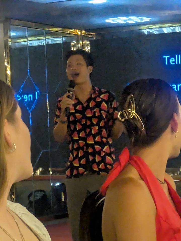 Indoor scene of a host singing into a microphone wearing a watermelon-print shirt during evening entertainment.