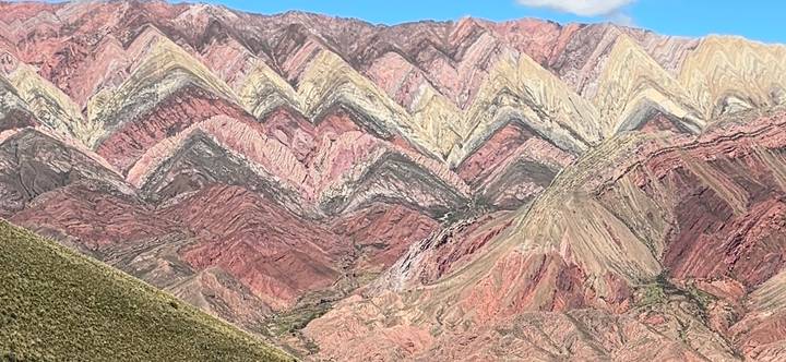 Multicolored zig-zag sedimentary mountain ridges rise beneath a blue sky.