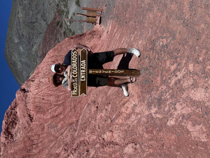 A couple poses beside a wooden sign for Paseo de los Colorados with red rocky hills behind.