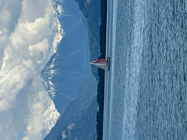 Single sailboat gliding across a blue alpine lake under towering snow-capped mountains and dramatic clouds.