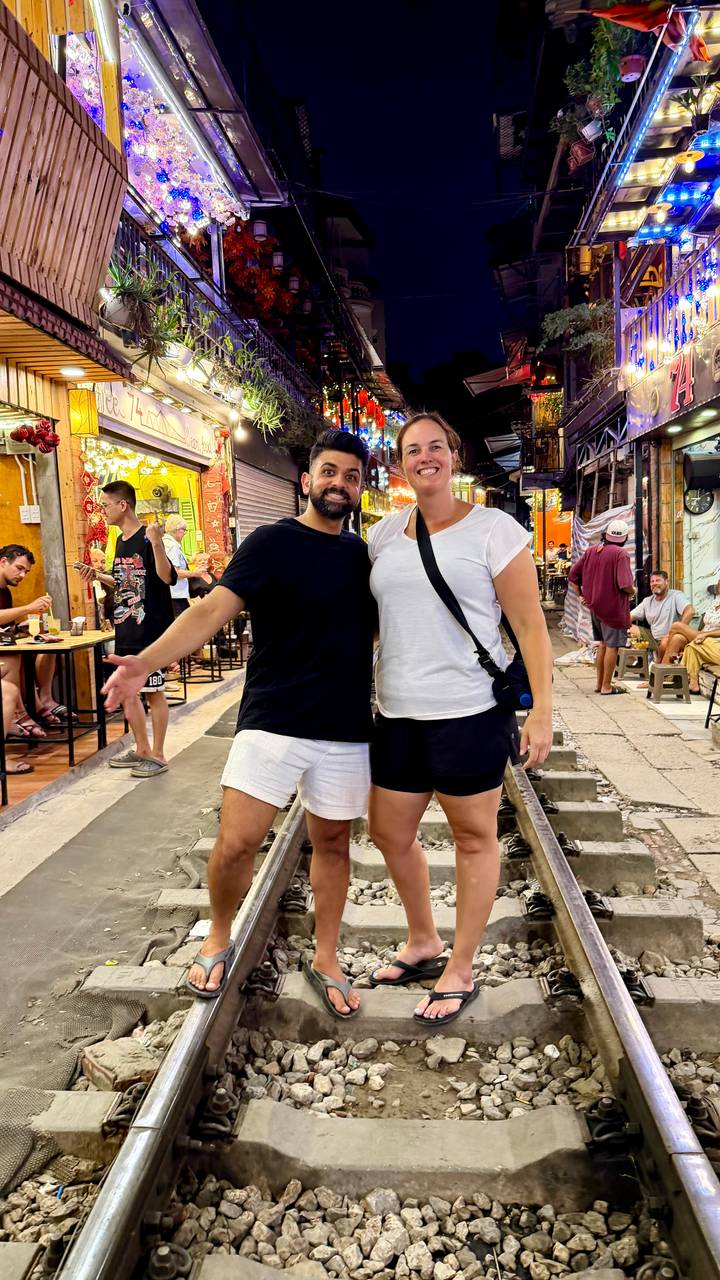 Two travellers pose on Hanoi’s famous Train Street at night, colourful cafés lining the tracks.