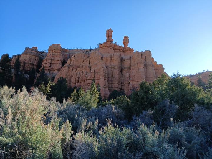 Sunlit red sandstone spires rise above juniper and sagebrush against a clear blue sky.