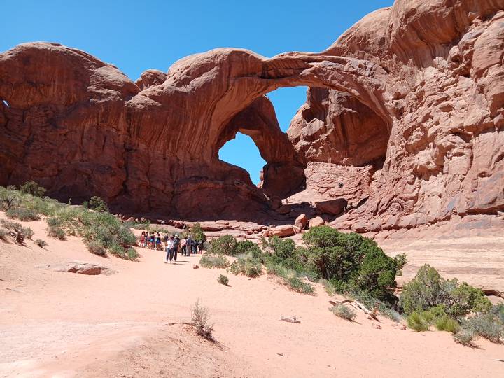Visitors approach the massive twin sandstone arches soaring above a sandy trail in bright sunshine.