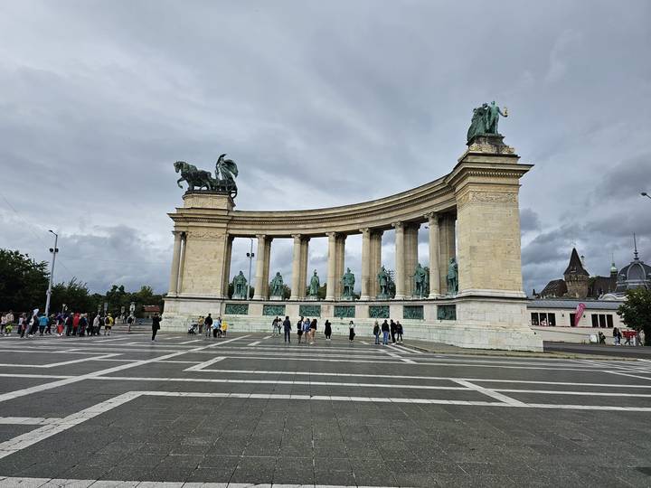 Wide plaza view of a grand semicircular colonnade topped with statues, with many tourists milling about.