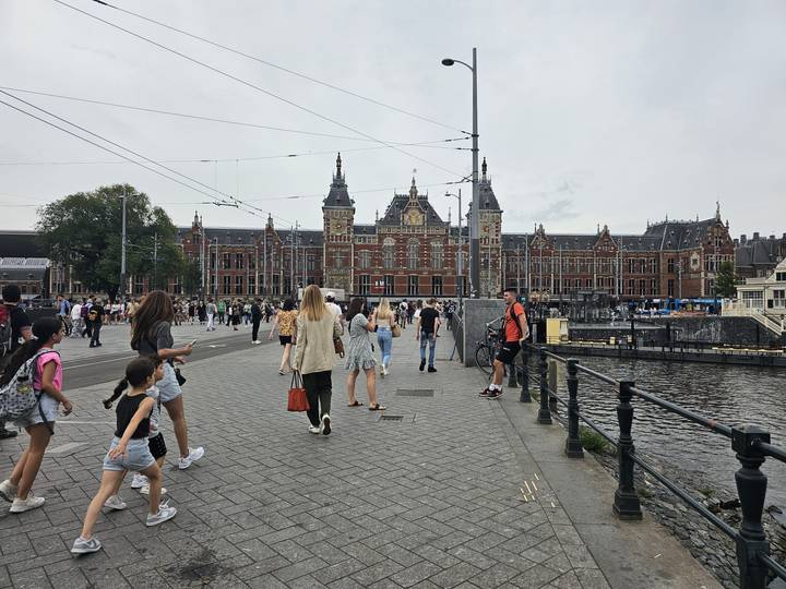 Busy waterfront scene with people walking in front of a historic red-brick railway station beside a canal.