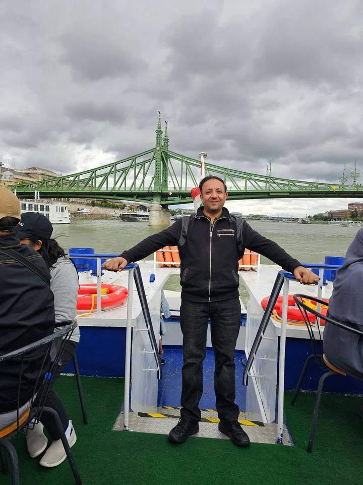 Man posing on the deck of a sightseeing boat with a green steel bridge and city skyline behind him.