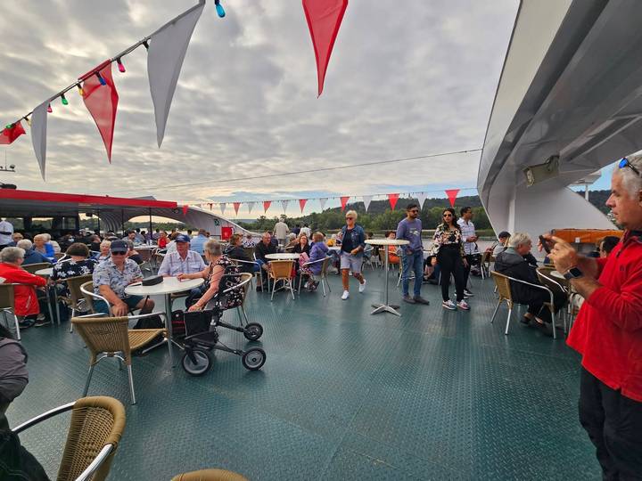Open deck of a river cruise ship with tables, bunting flags and passengers enjoying the view under a cloud-filled sky.