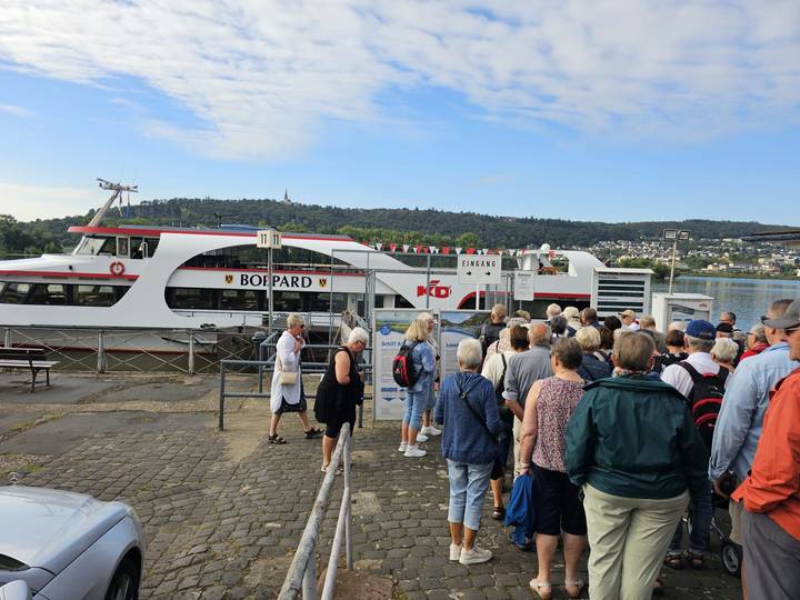 Line of tourists waiting to board a white excursion boat docked on a wide river.