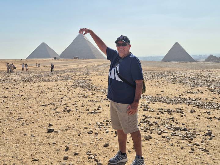 Tourist playfully posing to 'pinch' a pyramid at the Giza Plateau with multiple pyramids in view.