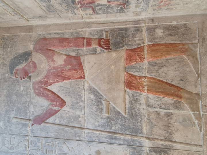 Ancient Egyptian wall relief of a standing figure holding a staff, with faded red and beige pigments.