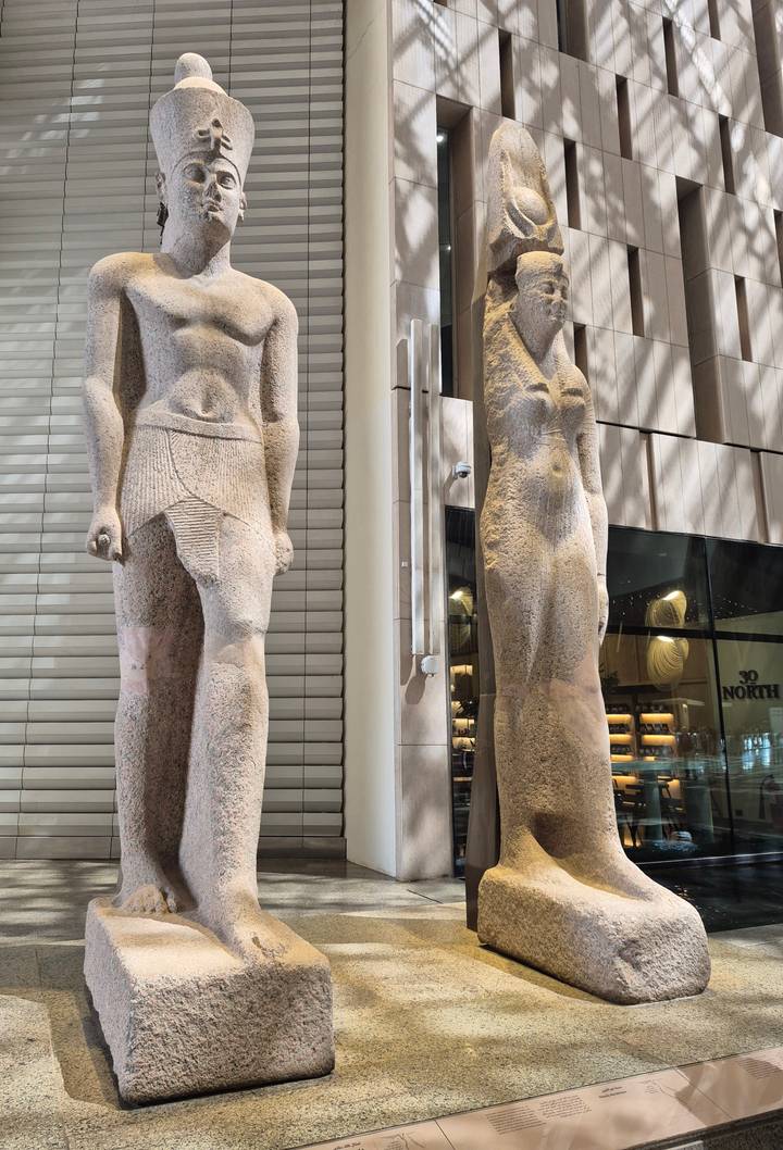 Two colossal ancient statues displayed upright inside a brightly lit modern museum atrium.