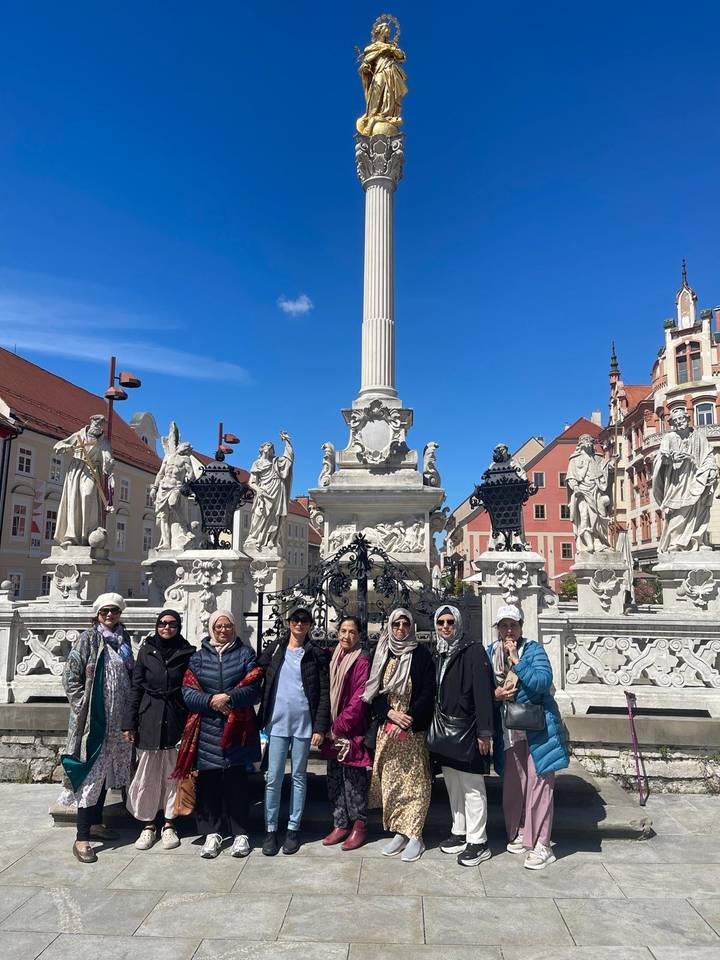 Tour group poses in front of ornate baroque column surrounded by stone statues in a European square
