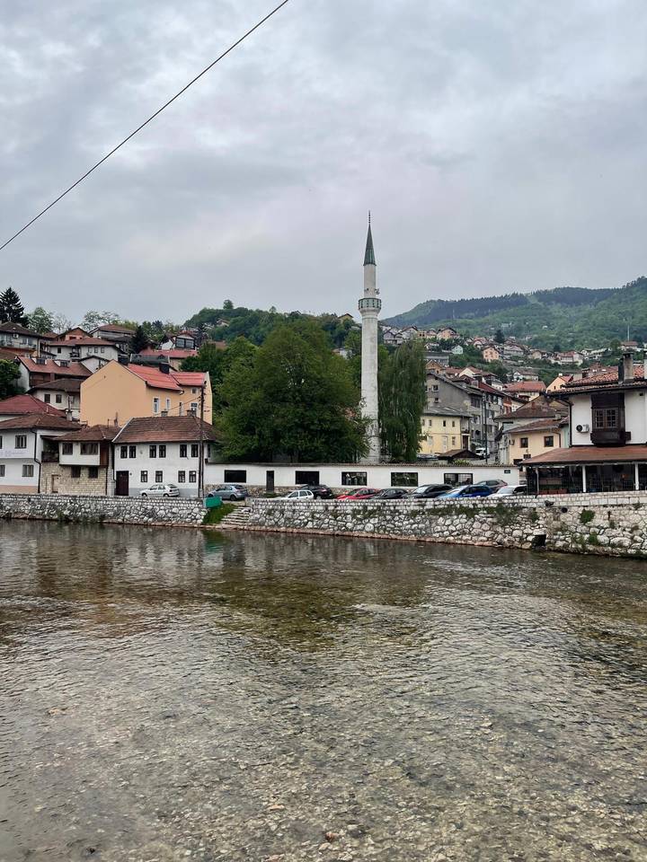 Riverbank view of Bosnian town with hillside houses and slender minaret under grey skies