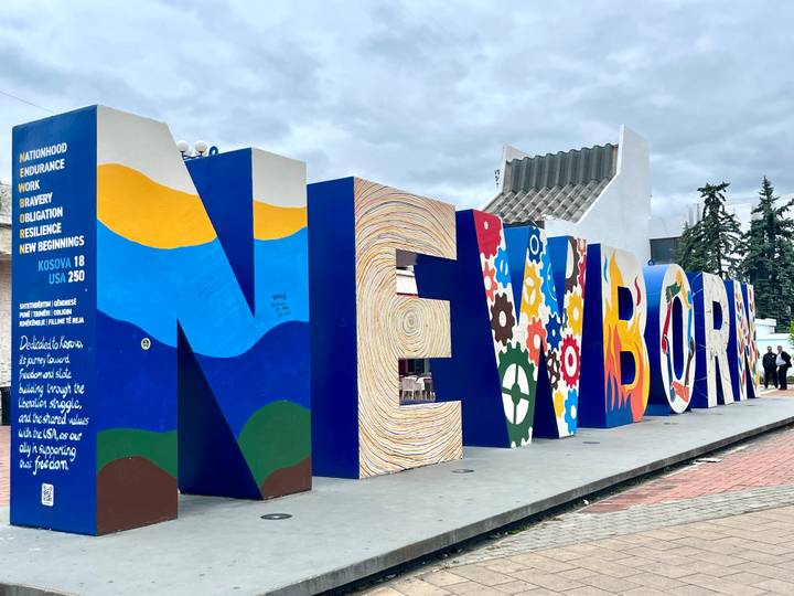 The colourful NEWBORN sculpture stands boldly on a plaza with a few people nearby.