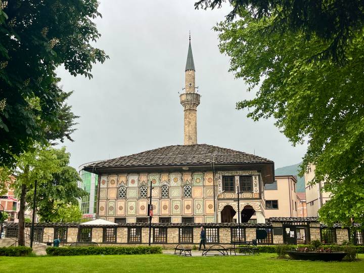A richly painted Ottoman-style mosque with a tall slender minaret framed by leafy trees on a grey day.