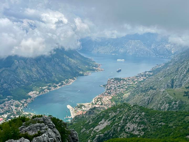 A sweeping aerial view of the Bay of Kotor snakes between dramatic mountains beneath drifting clouds.