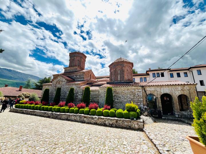 A stone monastery complex with manicured shrubs and bright flowers sits under a dramatic sky.