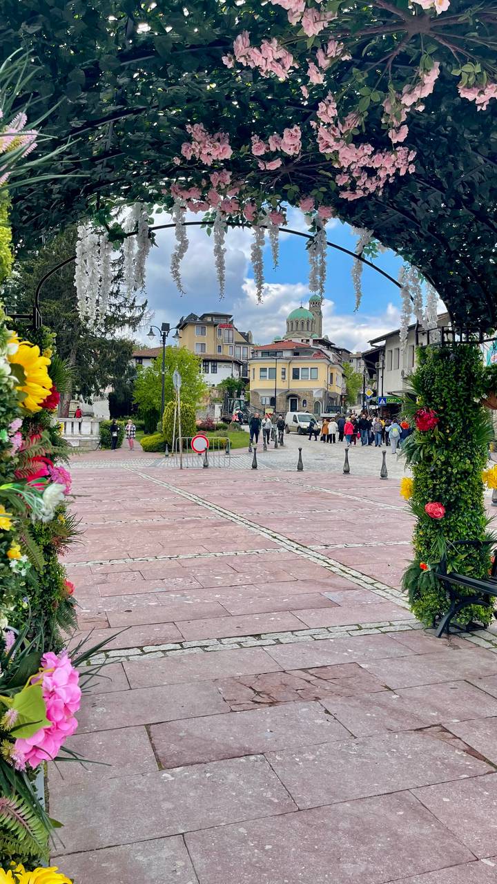 A lively town square framed by flower garlands bustles with people under a distant green-domed church.