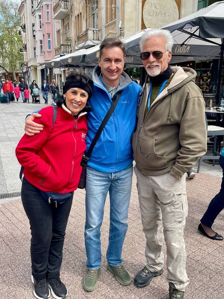 Three smiling friends pose arm-in-arm on a pedestrian street filled with cafés and shoppers.